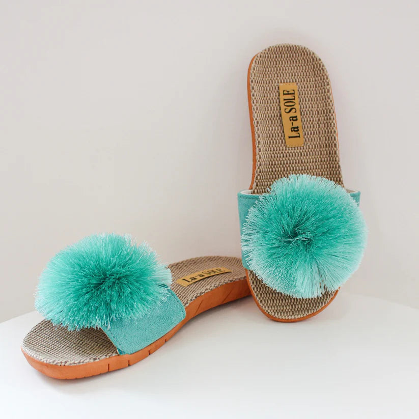A pair of aqua blue pom pom slides with a rubber slip-resistant sole and built-in arch for support, accompanied by a matching aqua blue strap.