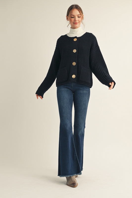 Woman wearing a navy cardigan with gold buttons, white turtleneck, and blue jeans on a beige background