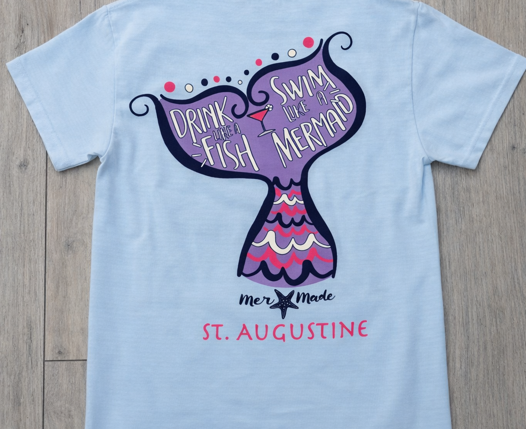 Back of light blue St. Augustine t-shirt with mermaid tail graphic and “Drink Like a Fish, Swim Like a Mermaid” text in pink, purple, and navy.