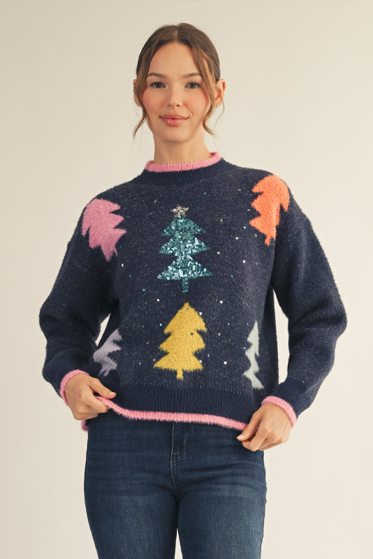 Model wearing a navy Christmas sweater with colorful tree patterns and a sparkling sequin tree in the center, paired with dark blue jeans — festive, cozy, and perfect for the holidays.