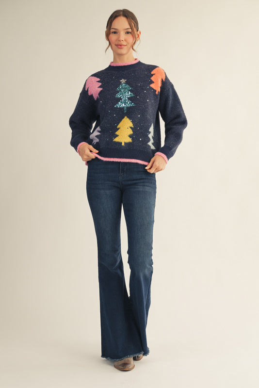 Model wearing a navy Christmas sweater with colorful tree patterns and a sparkling sequin tree in the center, paired with dark blue jeans — festive, cozy, and perfect for the holidays.