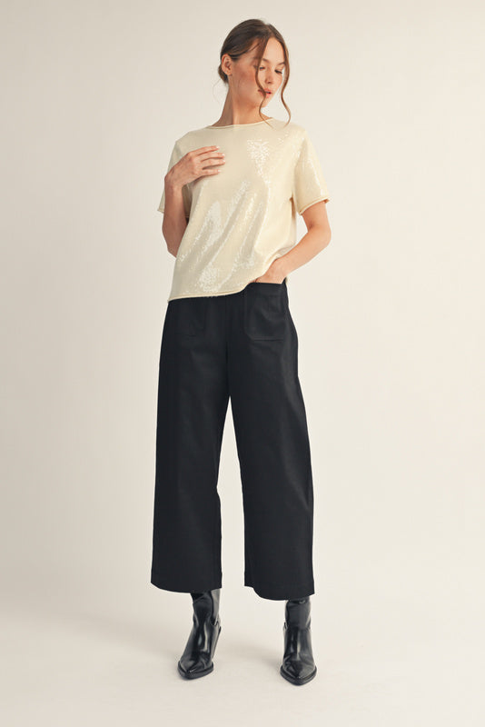 Model wearing a short-sleeve sequin top with a soft shimmer finish, paired with black wide-leg pants and ankle boots. The look is elegant, minimal, and perfect for a polished festive outfit.