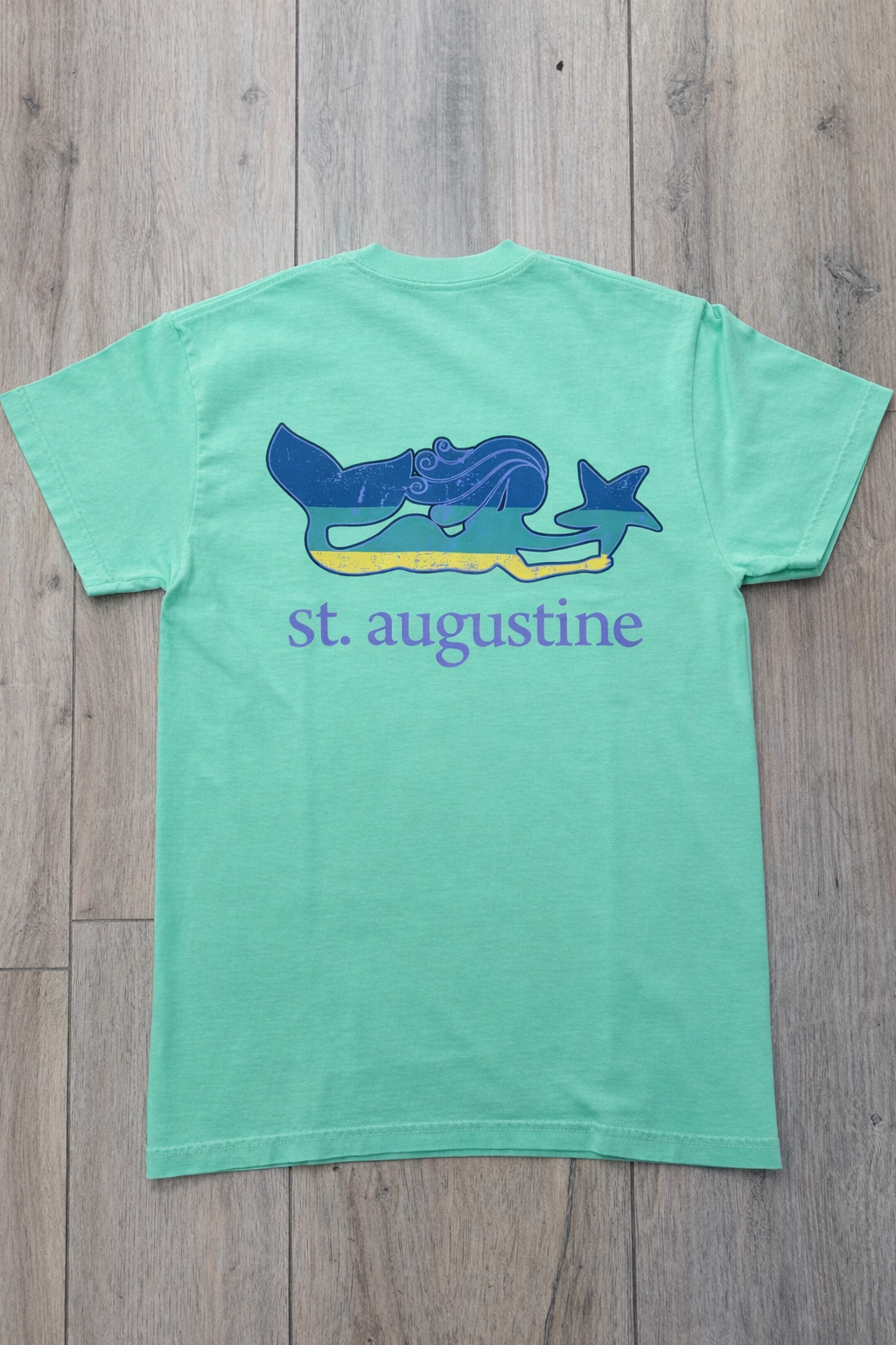 Back of mint green St. Augustine t-shirt with a stylized mermaid tail and starfish graphic above lavender “st. augustine” text.