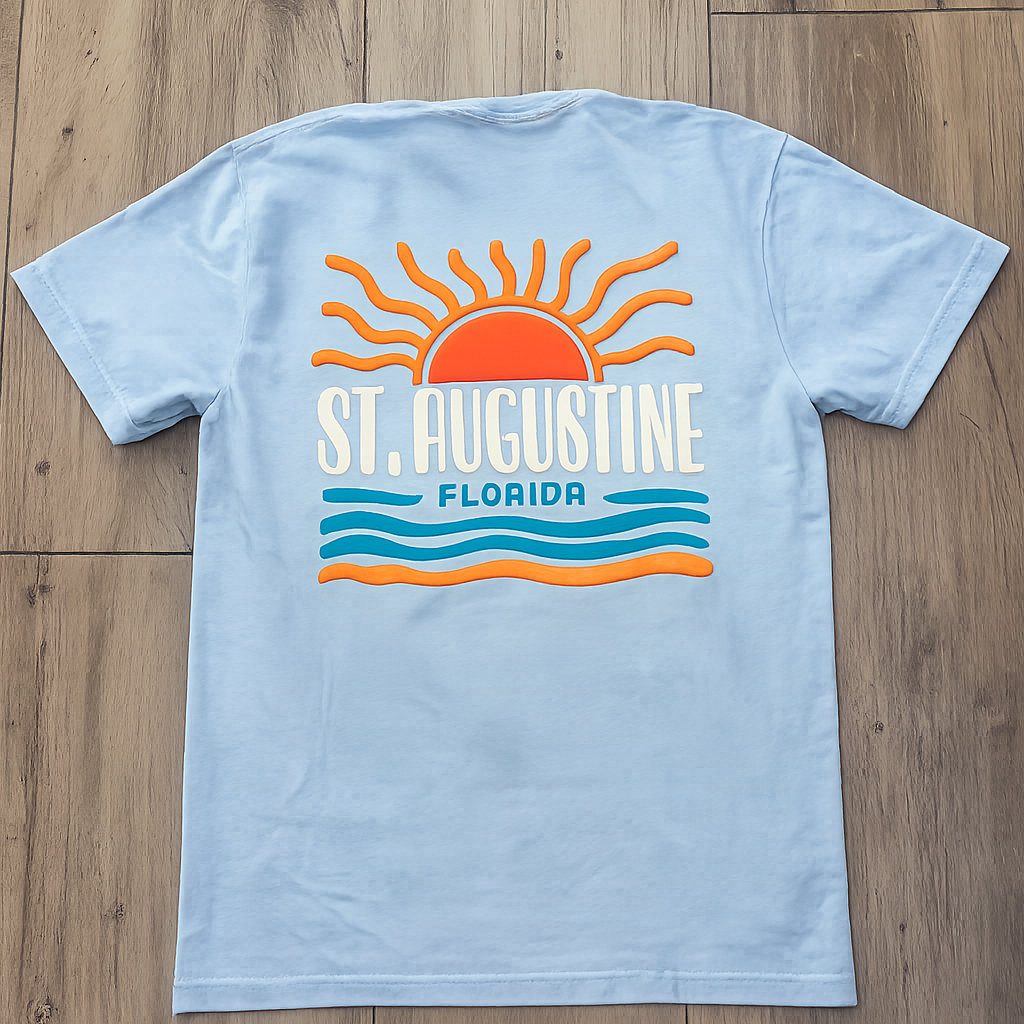 Back of light blue St. Augustine Florida t-shirt with orange sunrise graphic, white lettering, and layered blue and green wave stripes.