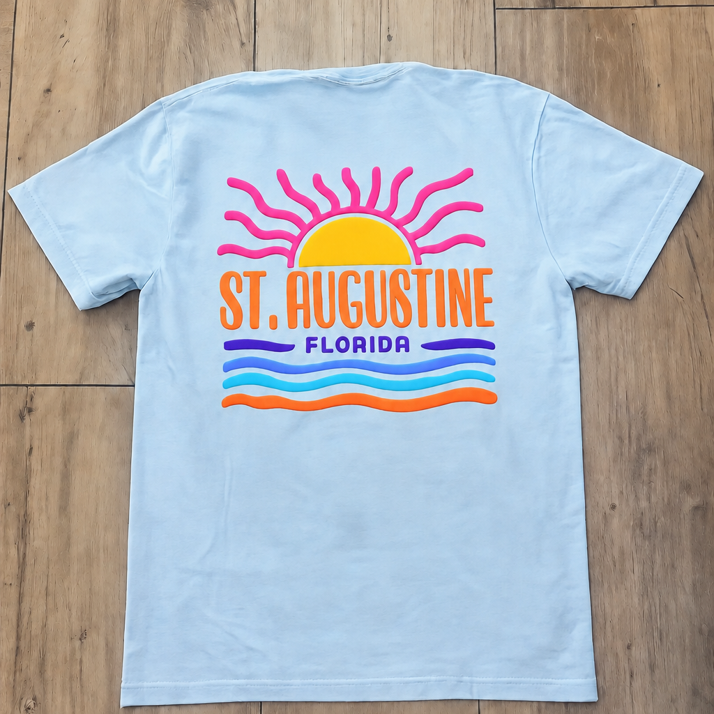Back of light blue St. Augustine Florida t-shirt with orange sunrise graphic, white lettering, and layered blue and green wave stripes.