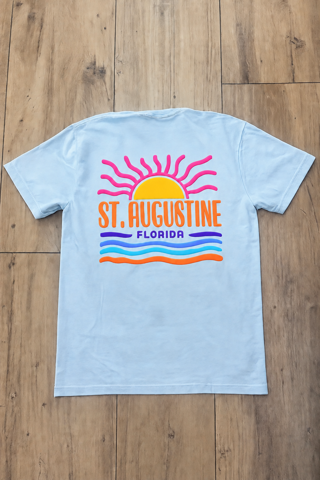 Back of light blue St. Augustine Florida t-shirt with orange sunrise graphic, white lettering, and layered blue and green wave stripes.