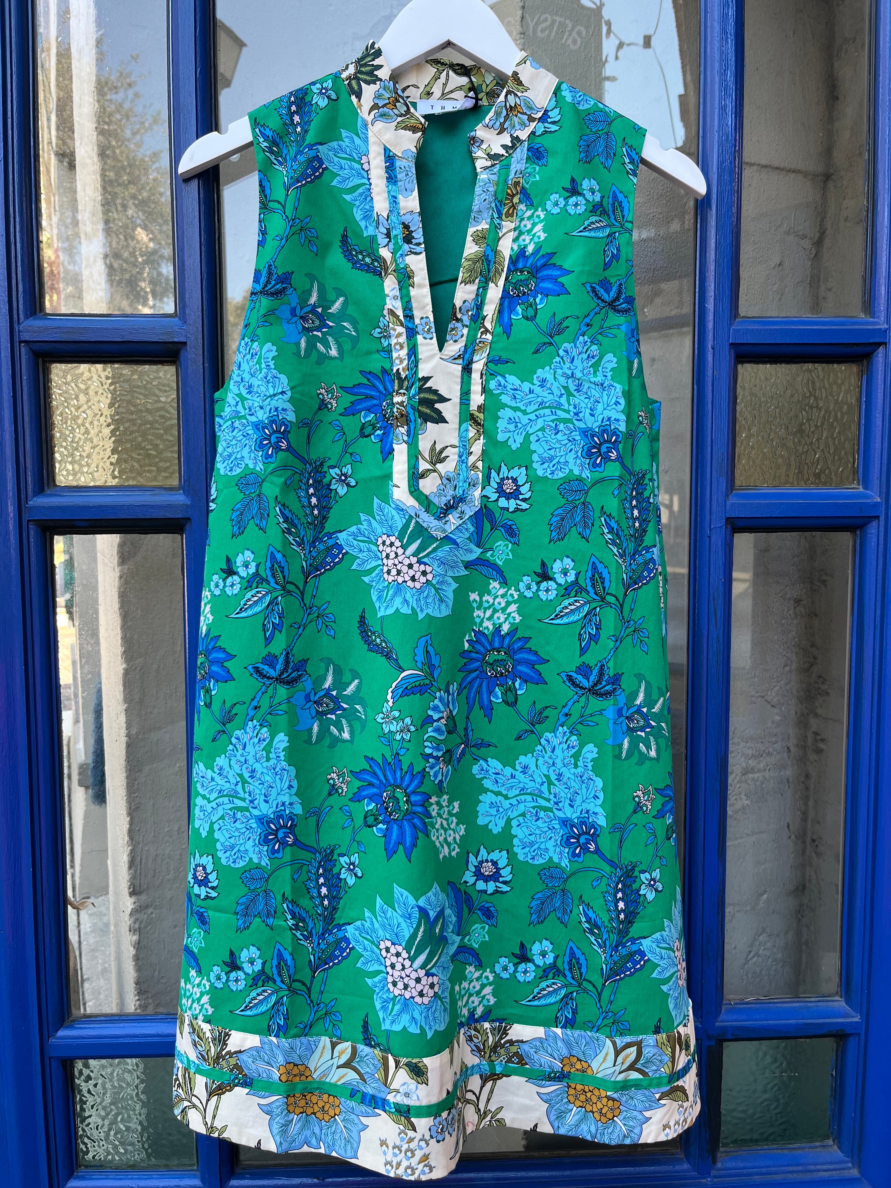 Sleeveless green dress with blue floral print, split V-neck collar with contrast floral trim, and a border-print hem, hanging on a white hanger.