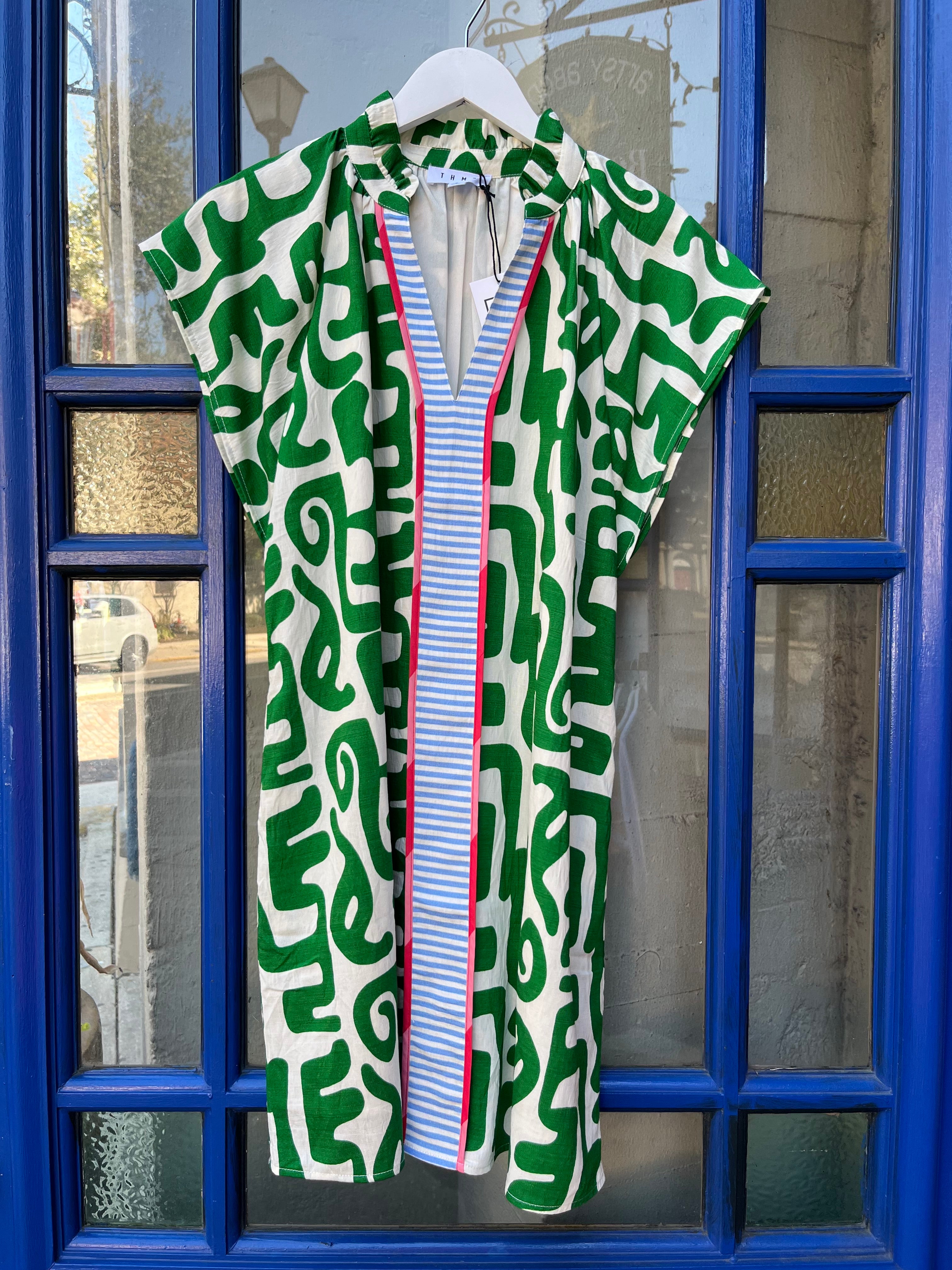 White caftan-style dress with bold green geometric print, split V-neck collar, and blue-and-white striped center placket with pink trim, hanging on a white hanger.