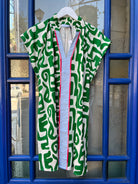 White caftan-style dress with bold green geometric print, split V-neck collar, and blue-and-white striped center placket with pink trim, hanging on a white hanger.