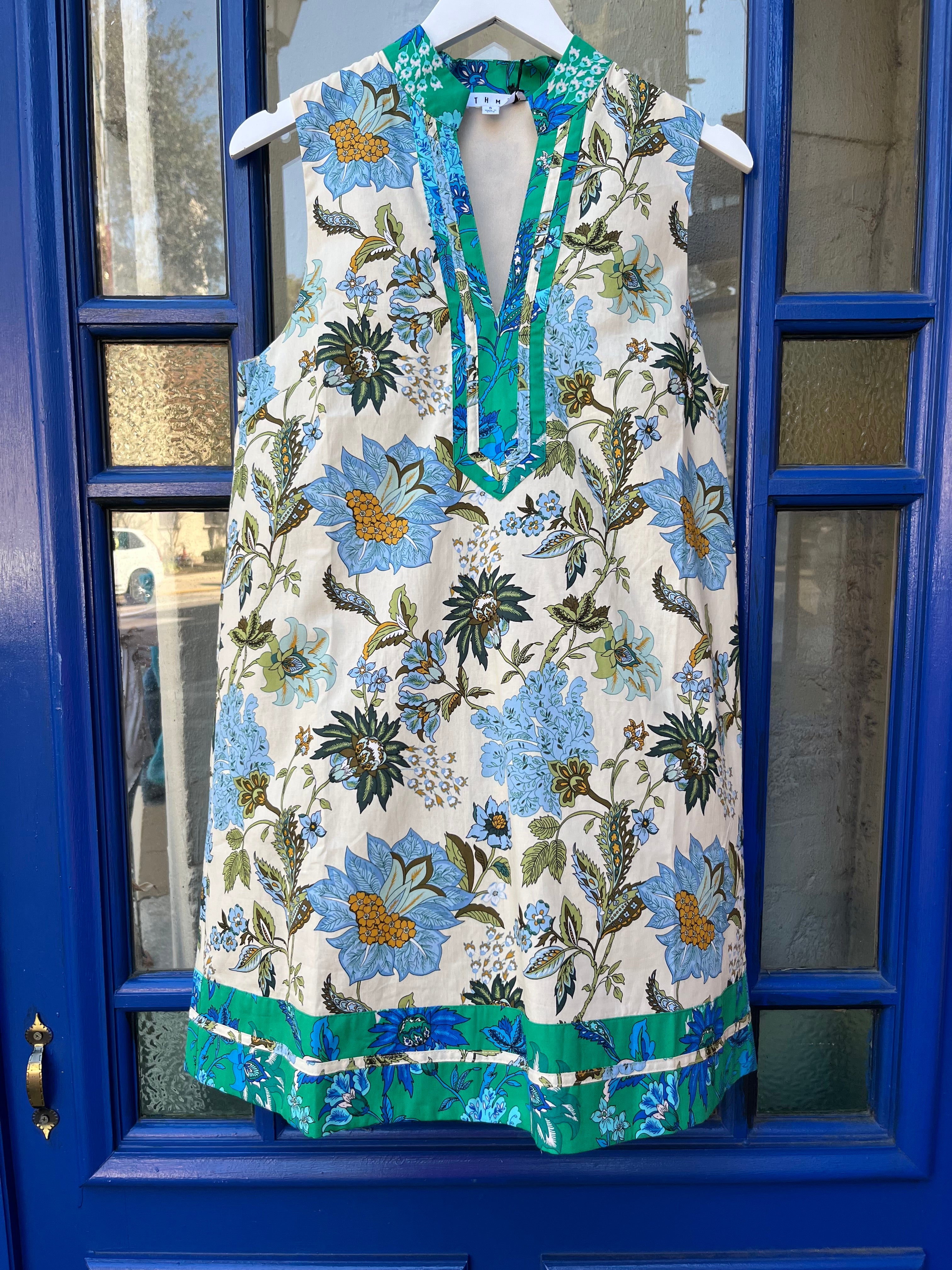 Sleeveless ivory shift dress with blue floral print, split V-neck collar with green contrast trim, and a green-and-blue border-print hem, hanging on a white hanger.