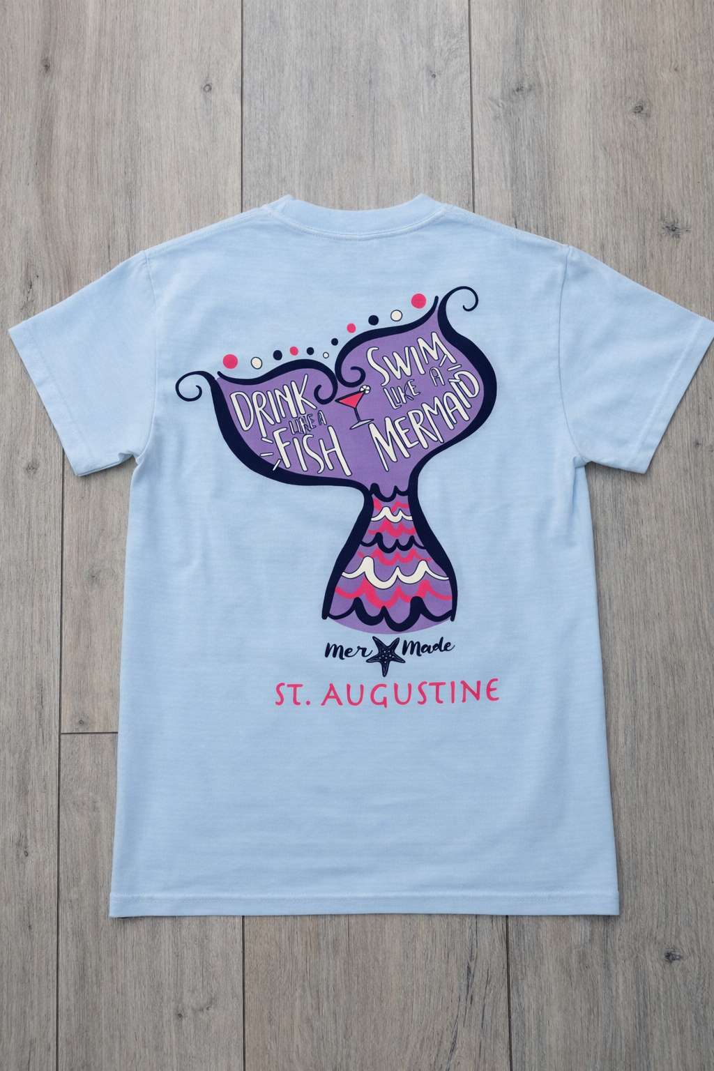 Back of light blue St. Augustine t-shirt with mermaid tail graphic and “Drink Like a Fish, Swim Like a Mermaid” text in pink, purple, and navy.