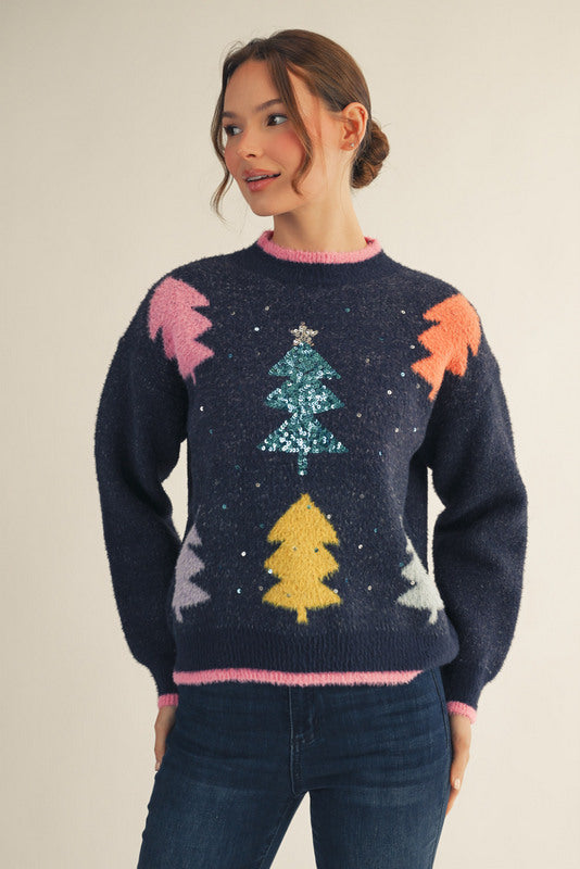 Model wearing a navy Christmas sweater with colorful tree patterns and a sparkling sequin tree in the center, paired with dark blue jeans — festive, cozy, and perfect for the holidays.