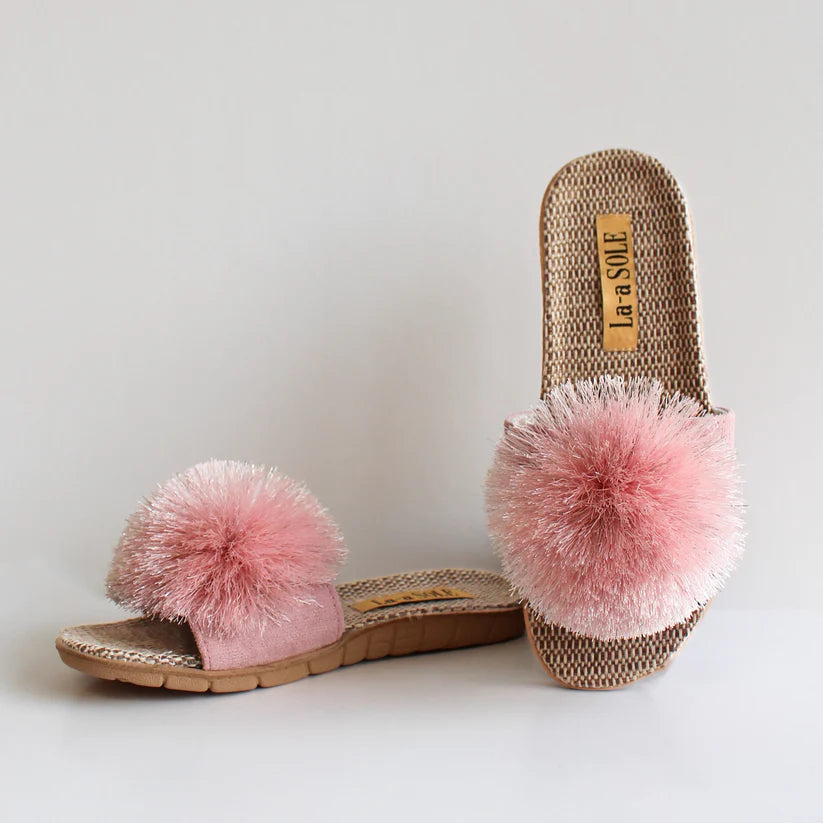 A pair of Pink Shimmer Pom Pom Slide Sandals featuring an oversized sparkling pink pom pom on the strap, with a lightweight woven sole and cushioned footbed, designed for playful coastal style.