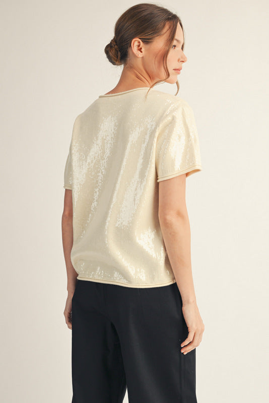 Model wearing a short-sleeve sequin top with a soft shimmer finish, paired with black wide-leg pants and ankle boots. The look is elegant, minimal, and perfect for a polished festive outfit.